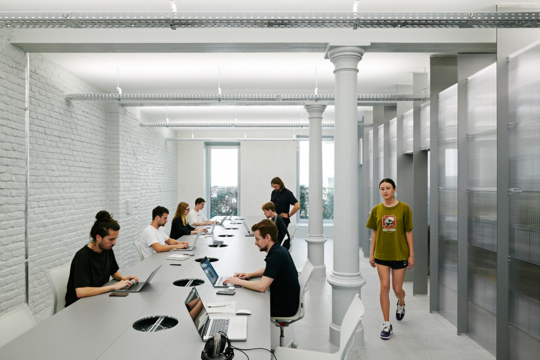 Bureau Borsche team members working at a long white desk in a minimalist office with white brick walls and grey columns.