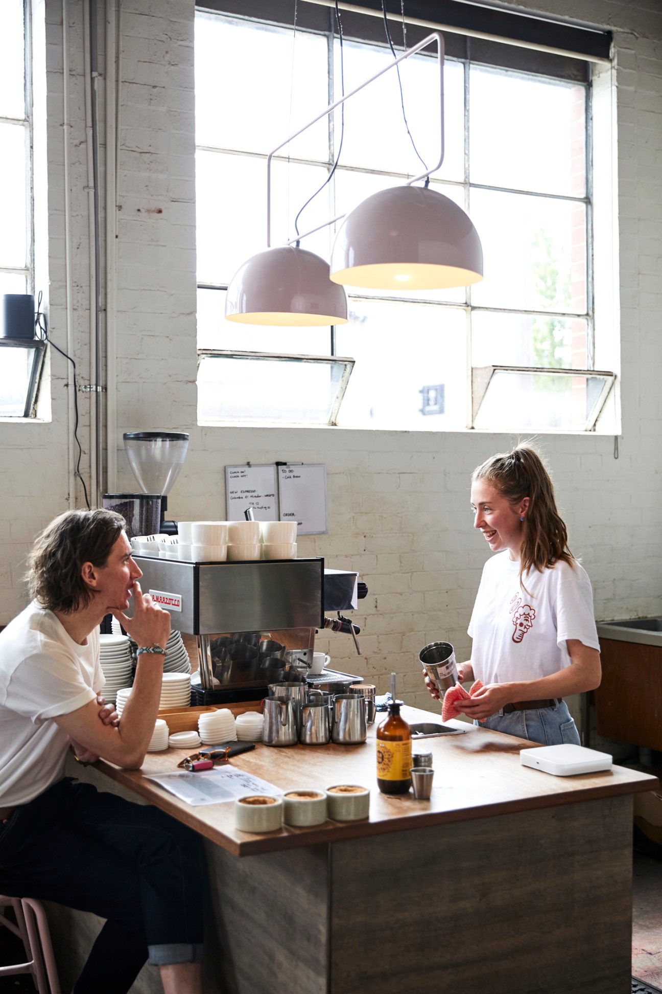 Two women chat over coffee in a bright cafe with pendant lamps and large windows in Girona.