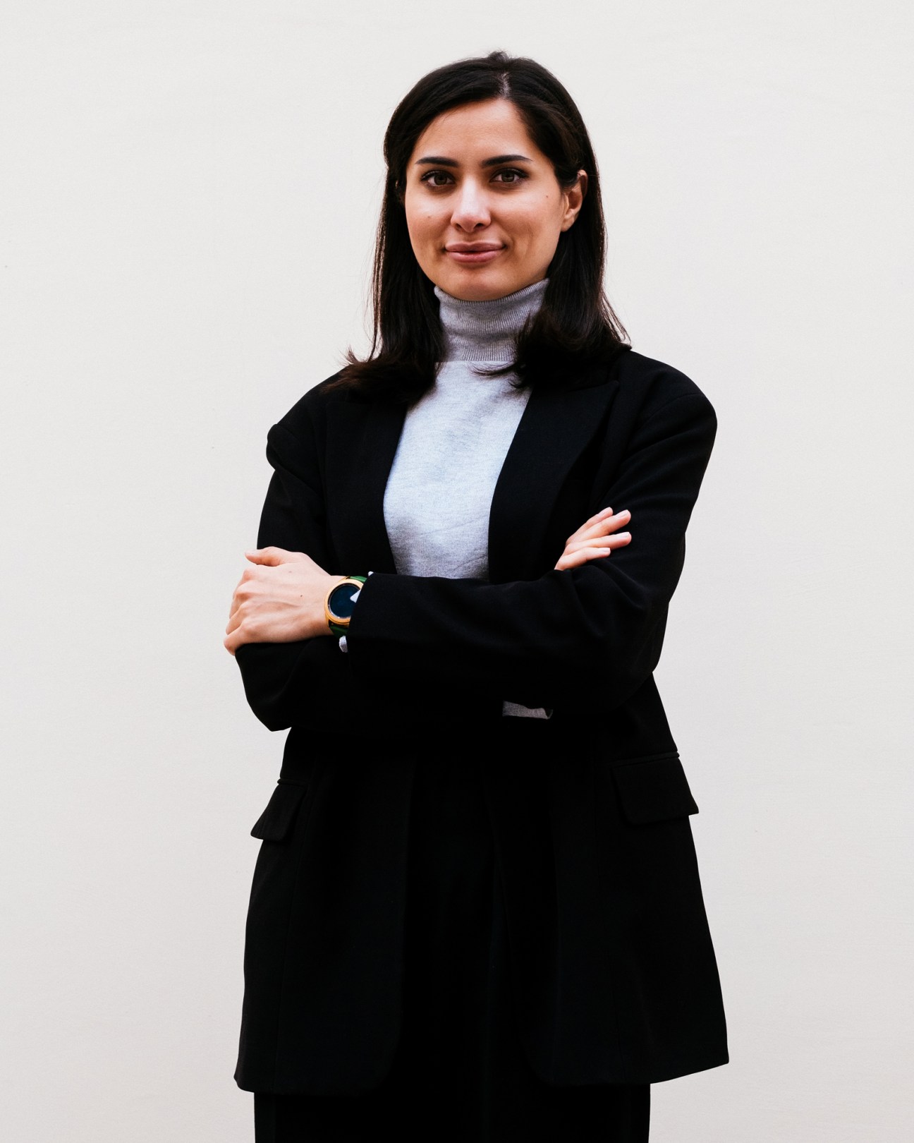 Madina Badalova wearing a black blazer and gray turtleneck with arms crossed, smiling at camera