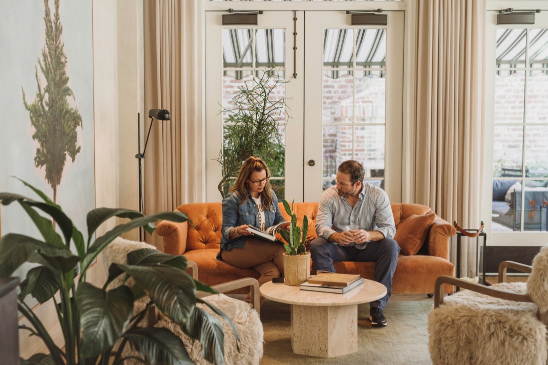 Man and woman sitting on orange velvet sofa in bright hotel room with large windows and plants