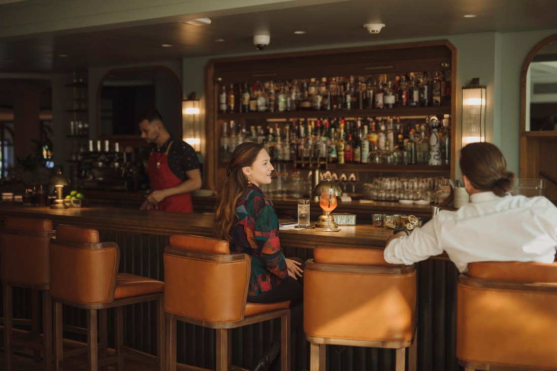 Bar area with orange leather stools, extensive bottle display, bartender and guests at Emeline hotel