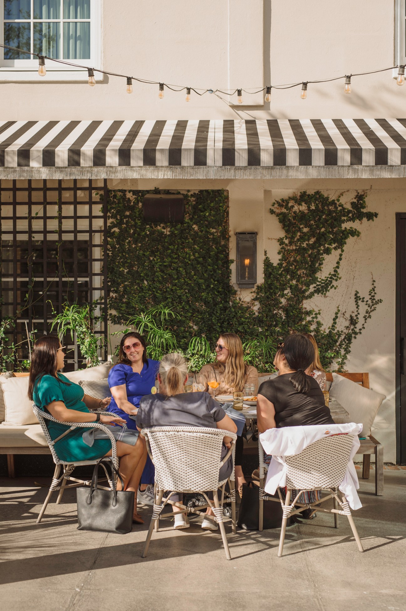 Group of women socializing at outdoor patio seating under striped awning with string lights