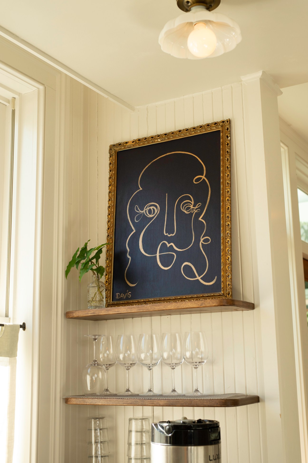Ornate framed artwork depicting a face in gold lines on dark background above shelves with wine glasses