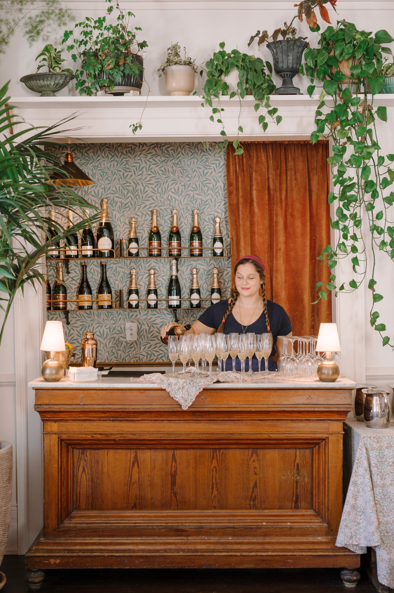 Woman in black top arranging wine glasses at wooden bar with bottles displayed on shelves and plants overhead