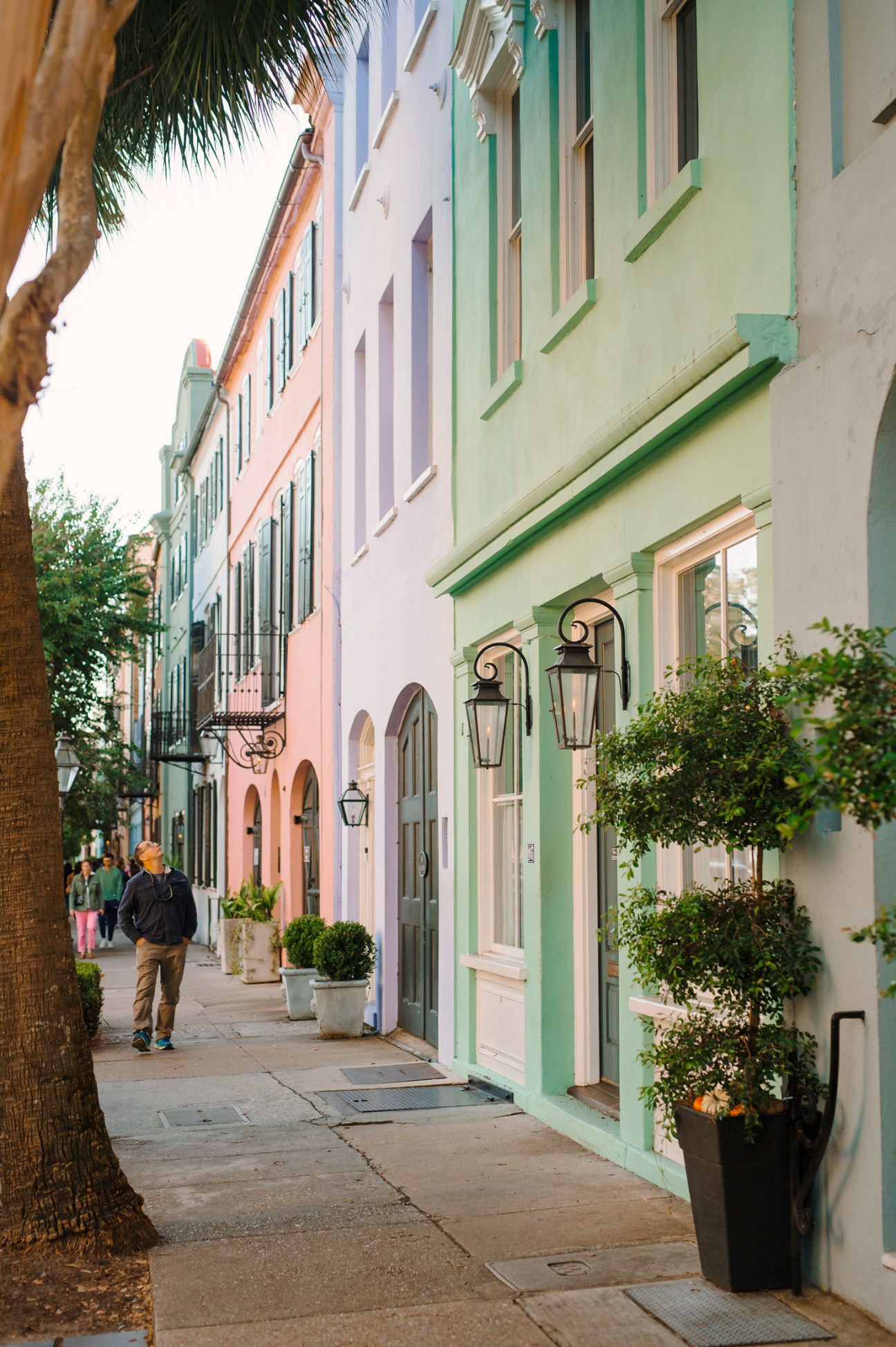 Pastel-colored historic buildings line a tree-shaded sidewalk in Charleston's downtown district