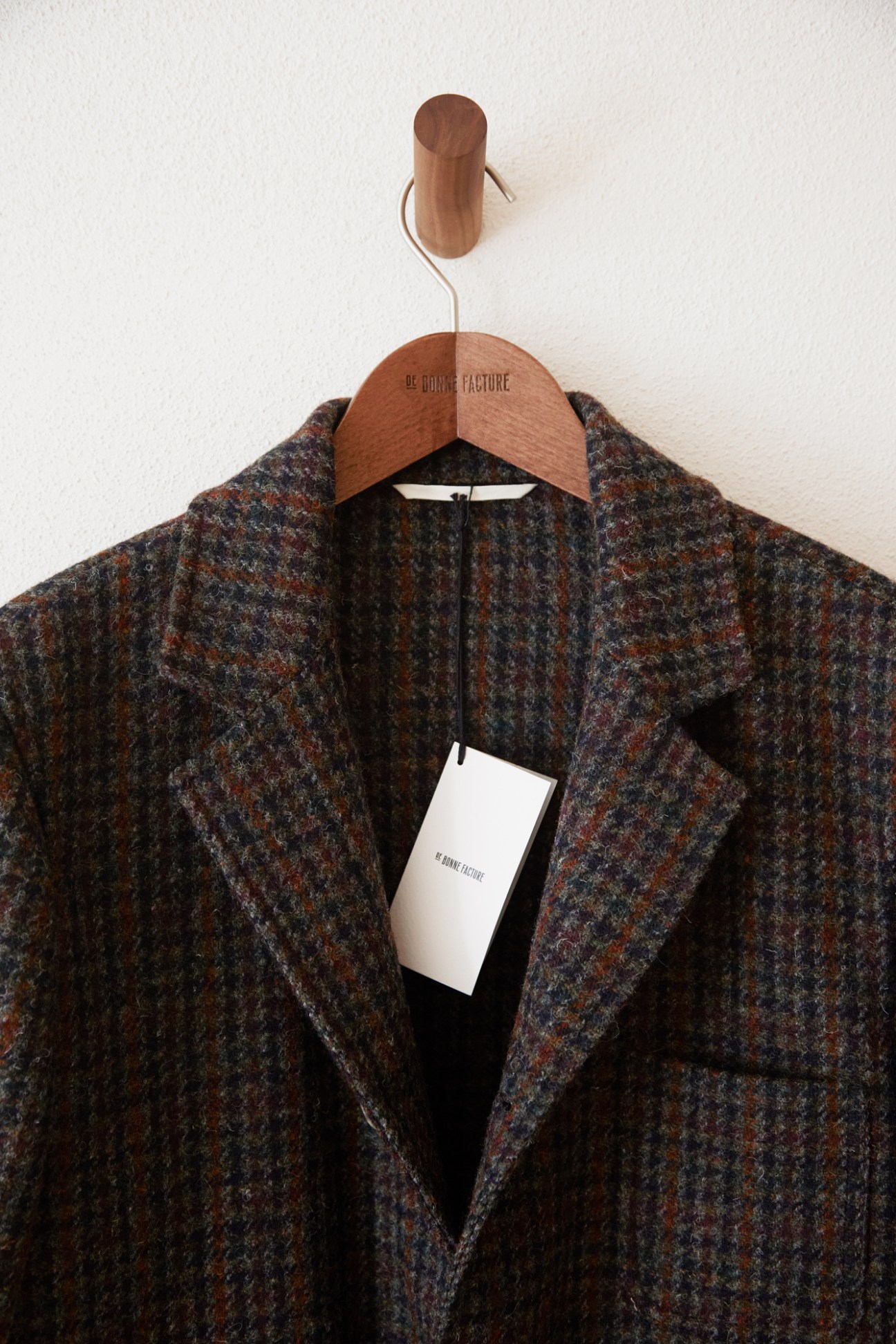 Tweed blazer hanging on wooden hanger with brand tag against white wall