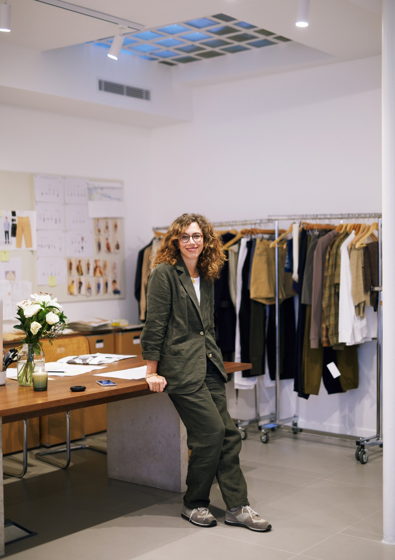 Woman with curly hair and glasses wearing green suit standing in modern retail space with clothing racks