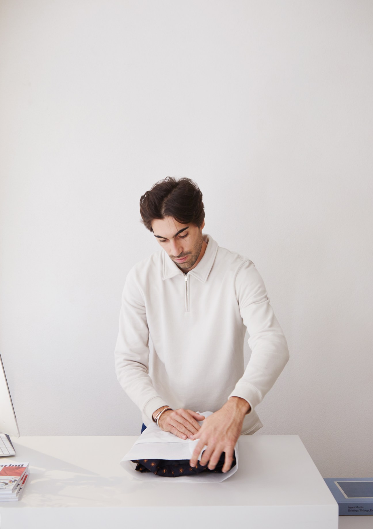 Person in white sweater carefully folding or examining a dark garment with small patterns on white surface