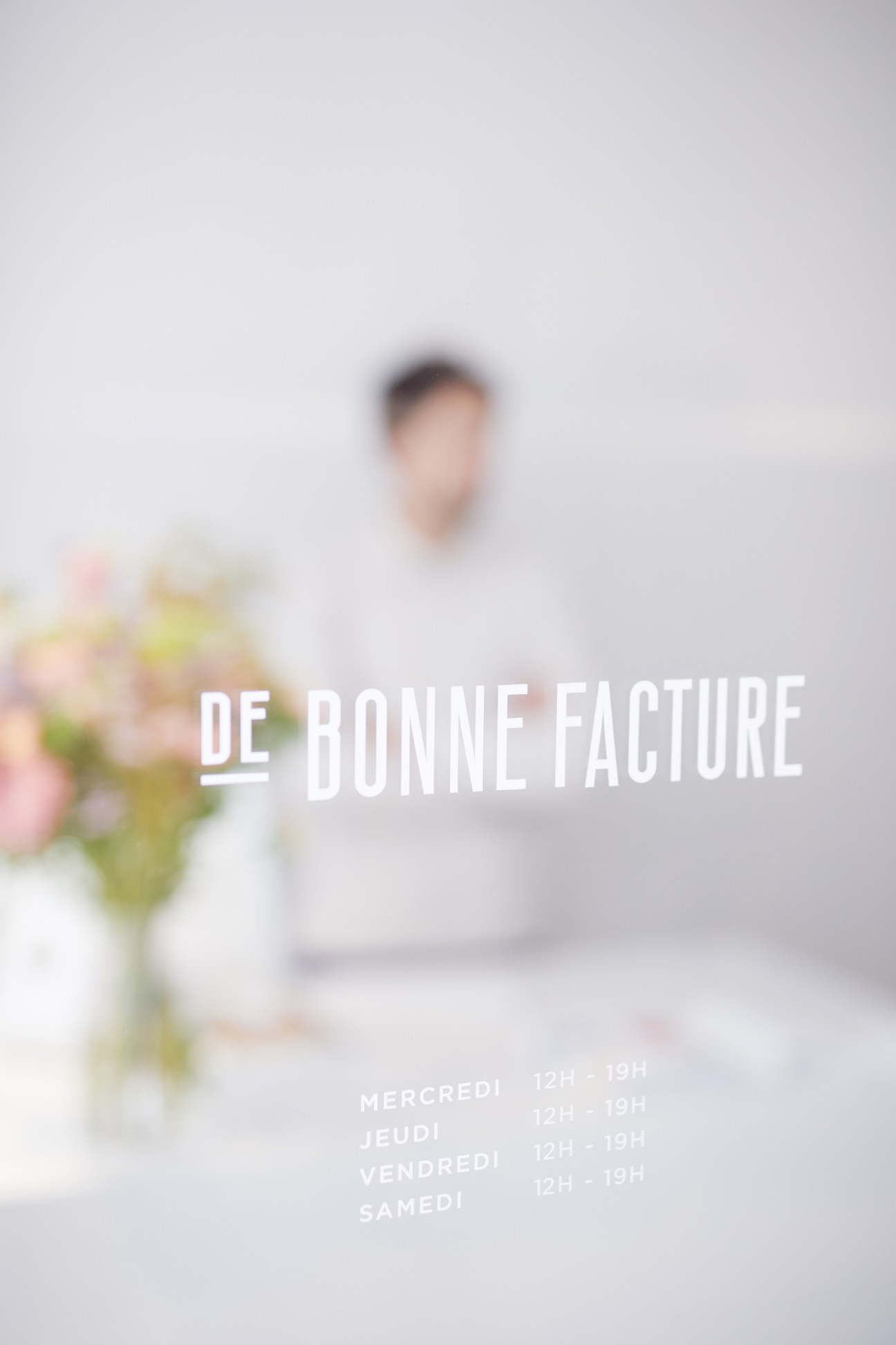 Blurred view through glass window showing De Bonne Facture store interior with person and flowers