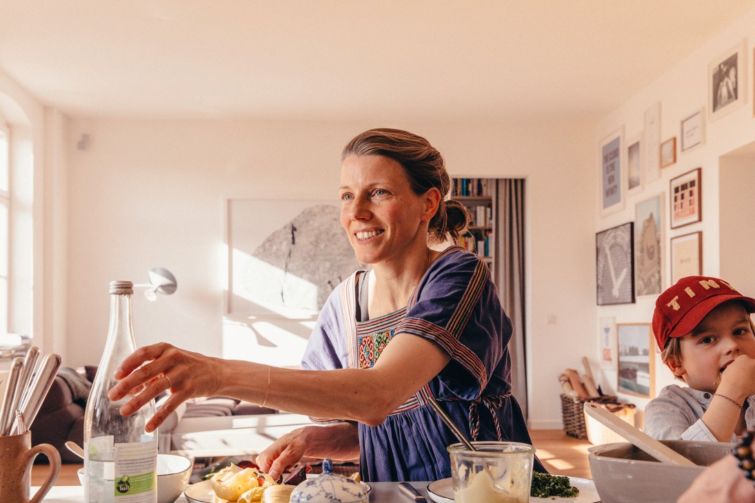 A smiling woman in a patterned top reaches across a dining table to pass a glass water bottle