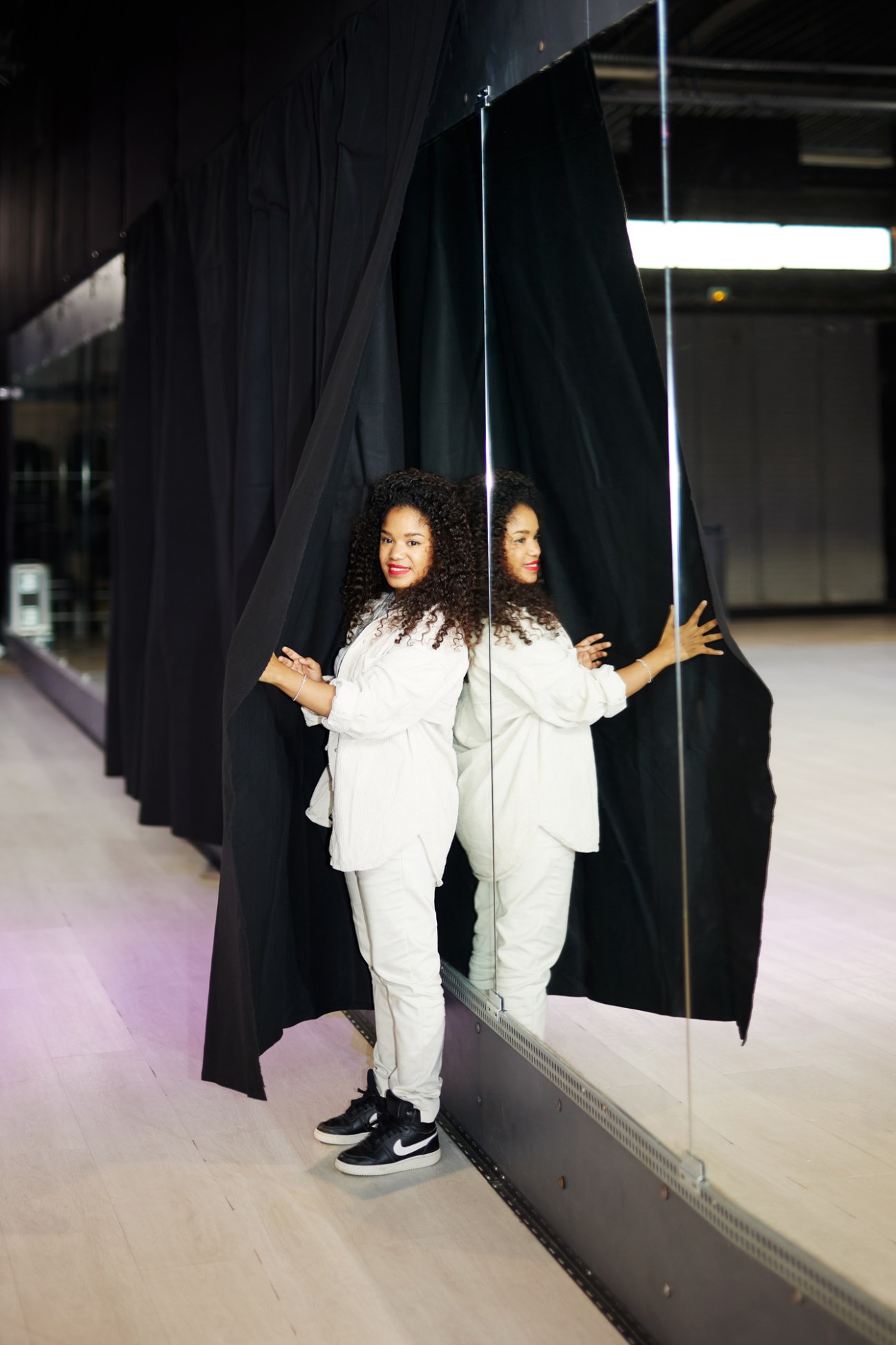 Woman with curly hair in white outfit standing against mirrored wall in modern interior space