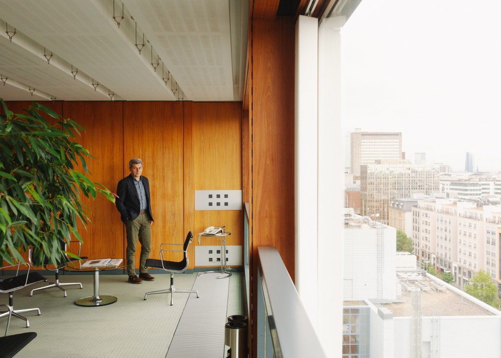 Man in dark blazer standing in modern office with wooden wall paneling and city view.