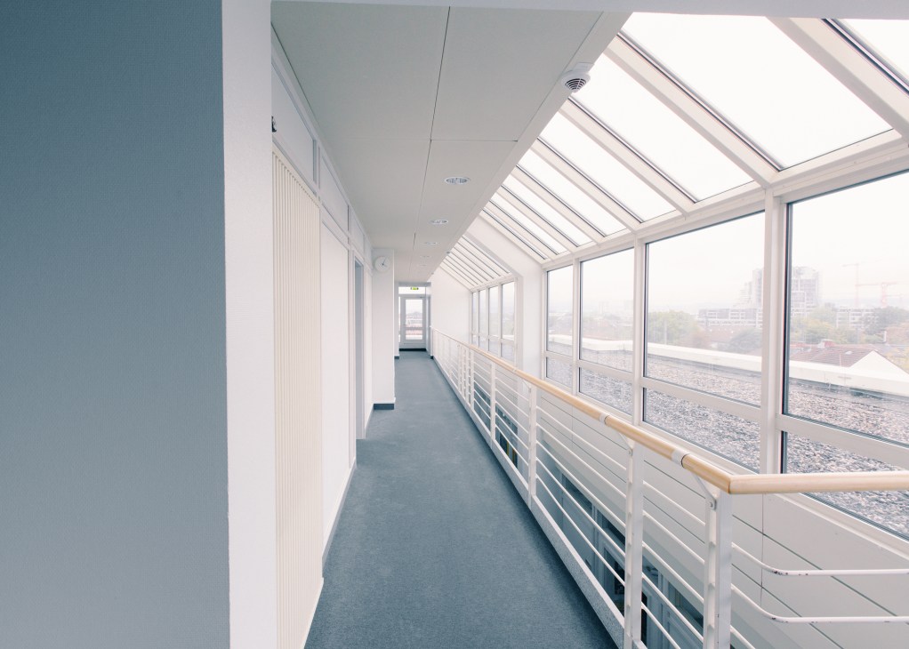 Modern office corridor with skylights, glass windows overlooking Frankfurt cityscape, wooden handrail, and gray flooring.