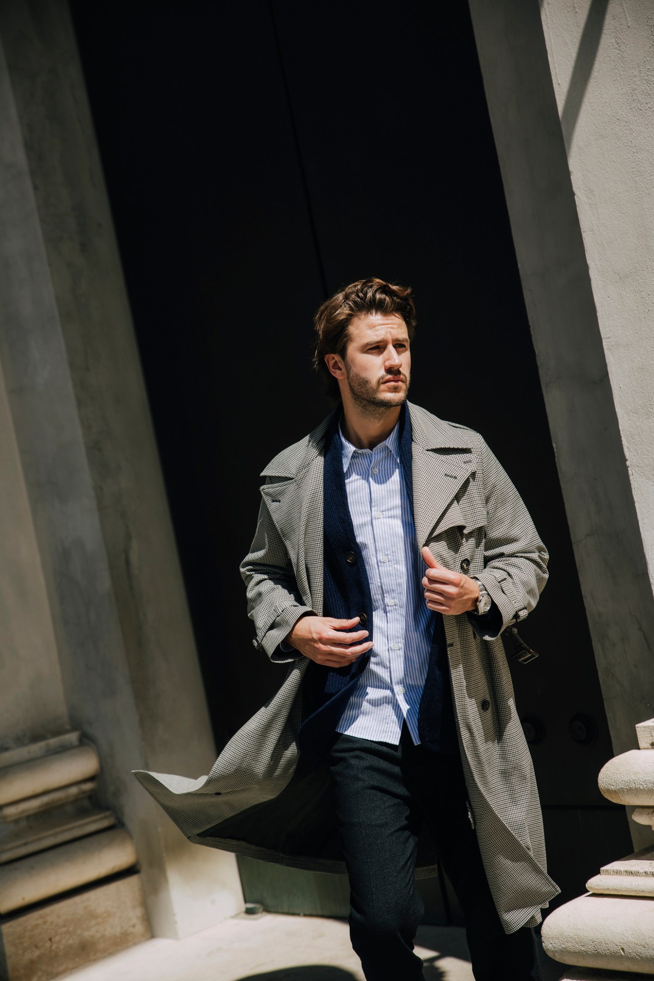Man in layered outfit with checkered trench coat and knit cardigan standing against modern concrete architecture