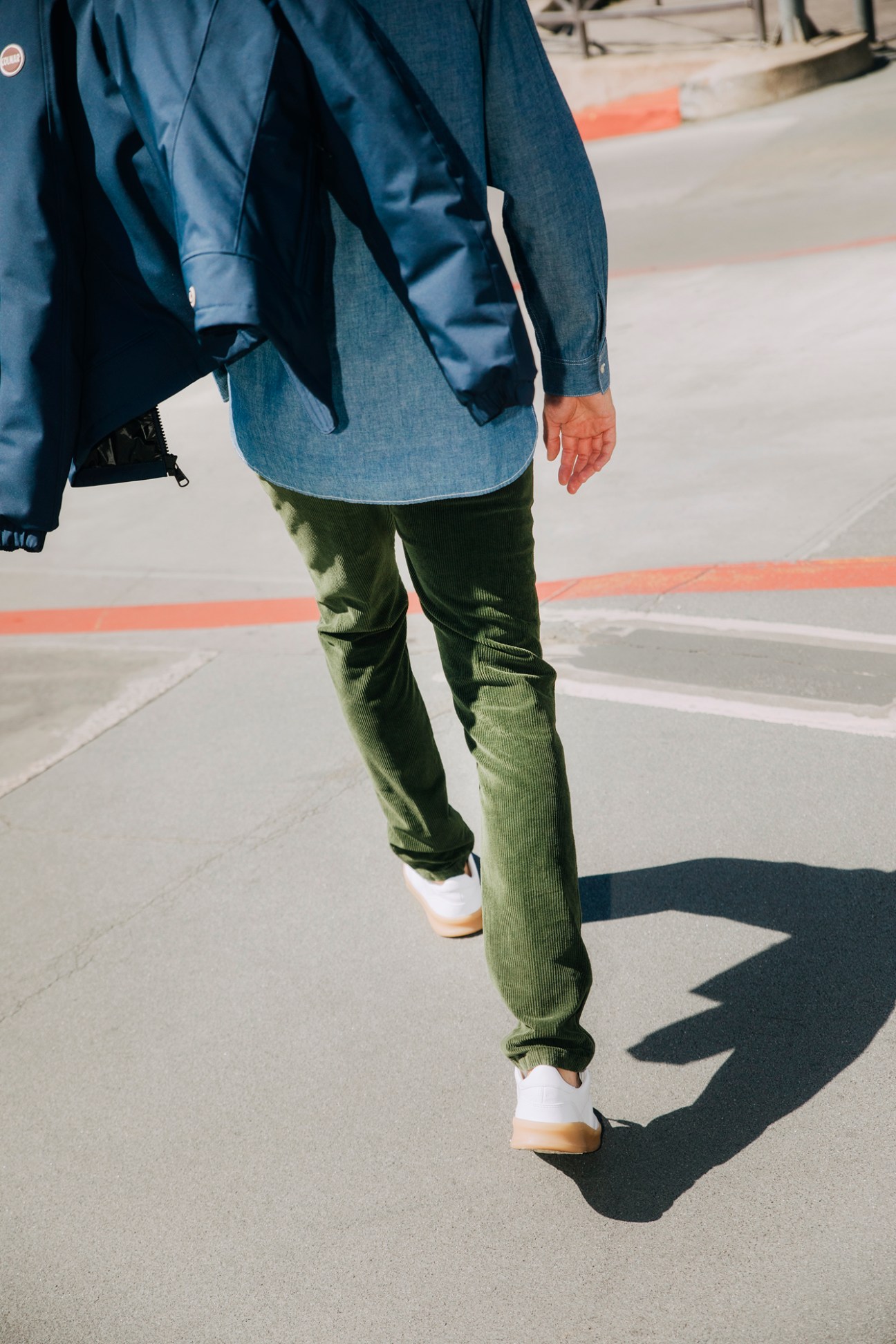 Person walking in green corduroy pants, blue denim jacket and white sneakers on concrete pavement with shadow visible
