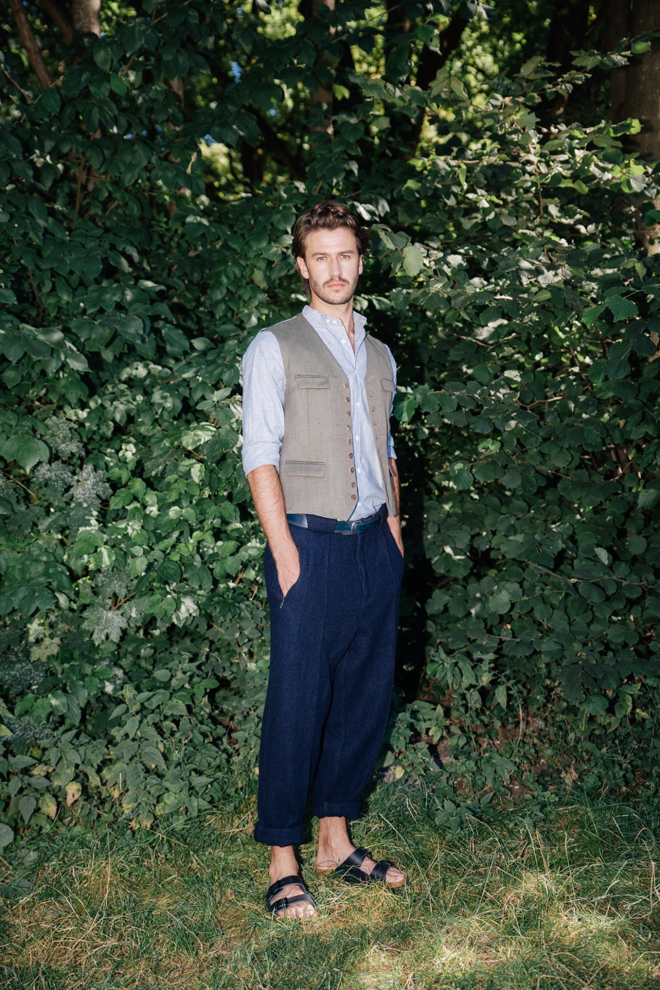 Male model in waistcoat, shirt, trousers and Birkenstock sandals standing against green foliage