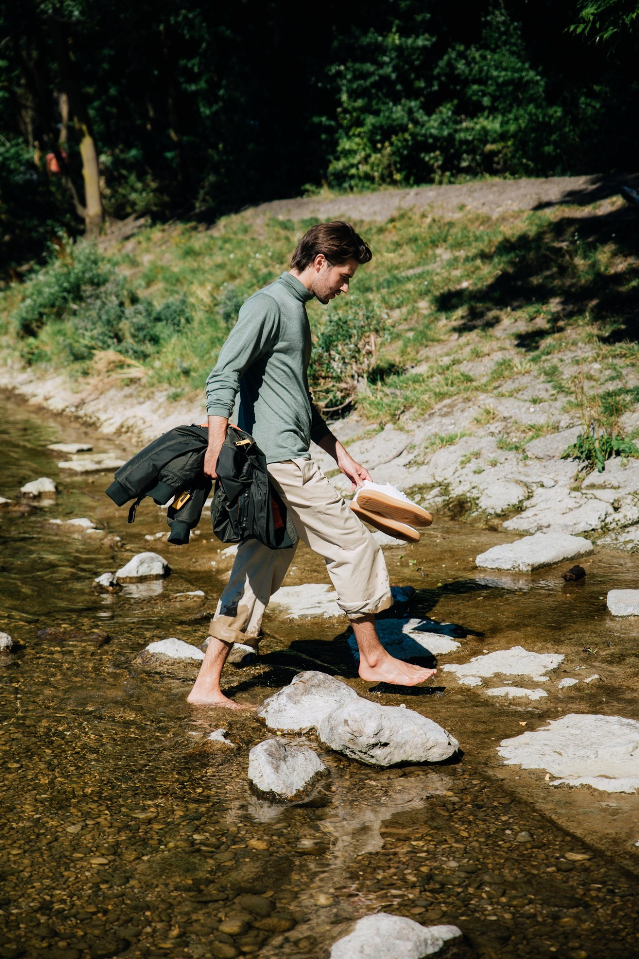 Man crossing shallow stream barefoot while carrying shoes and jacket, stepping carefully on rocks