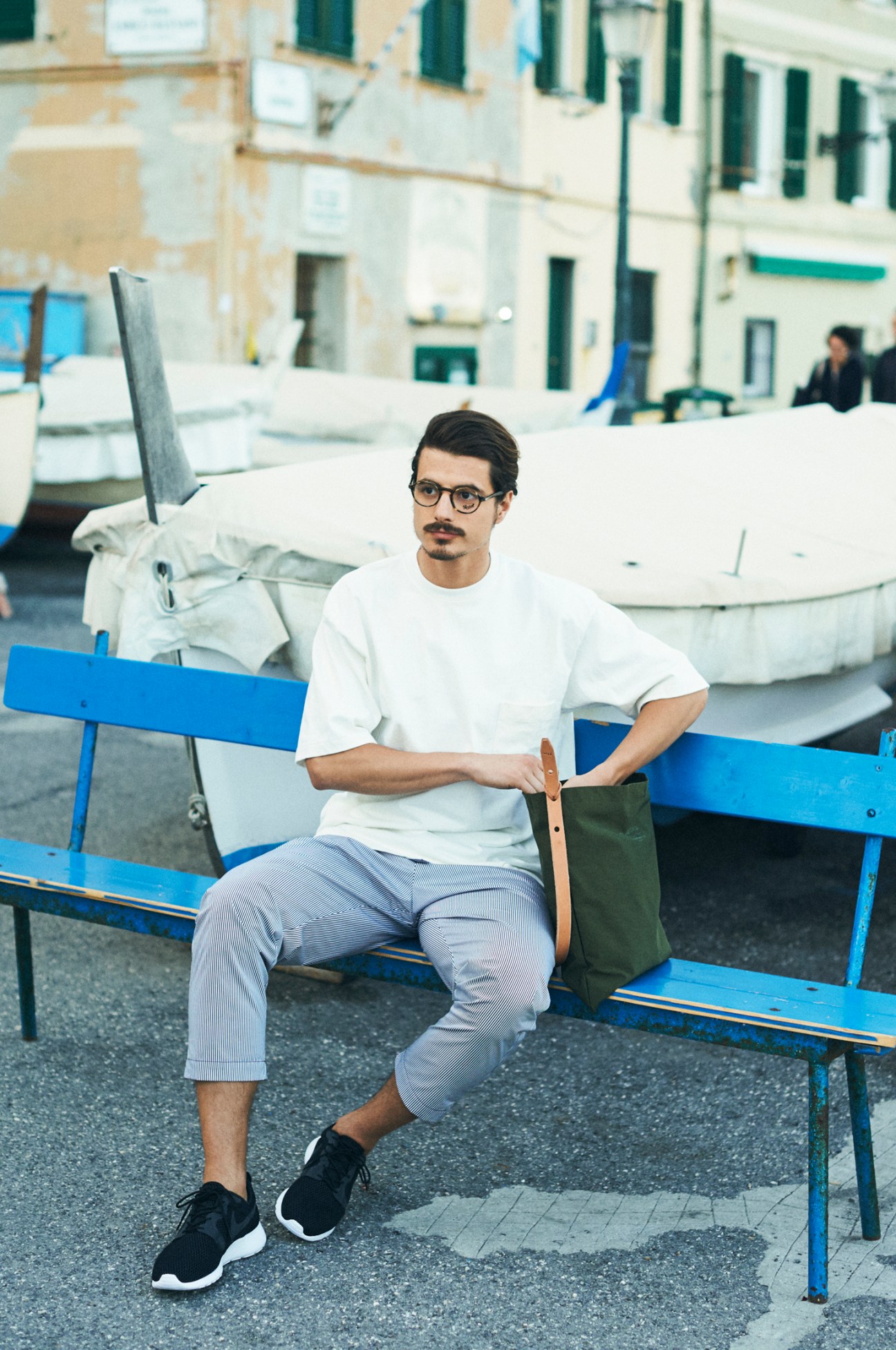 Man in white Auralee t-shirt and striped pants sitting on blue bench near boats in Mediterranean coastal town