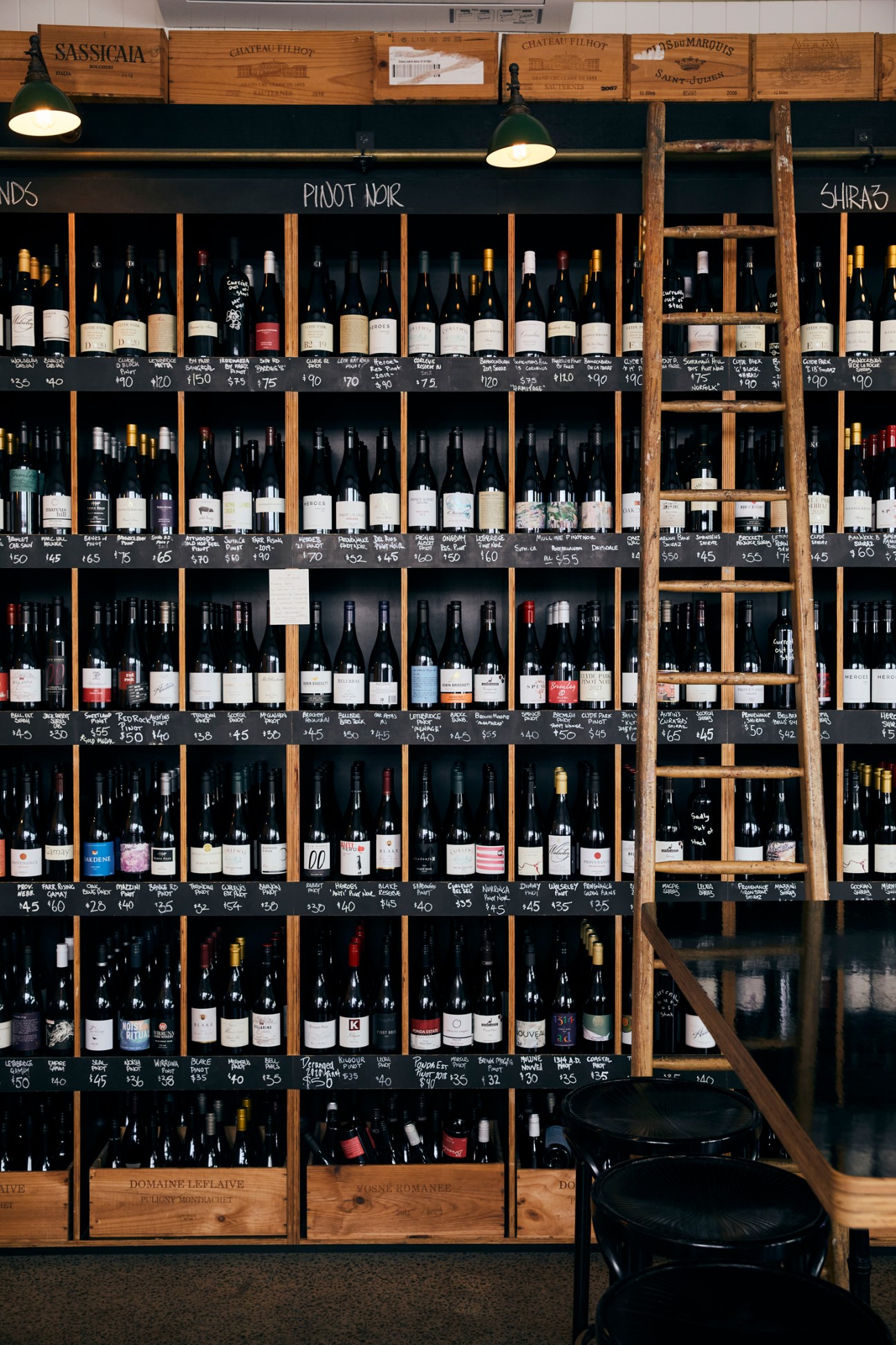Wine bottles displayed on wooden shelves with handwritten price labels in a wine shop.