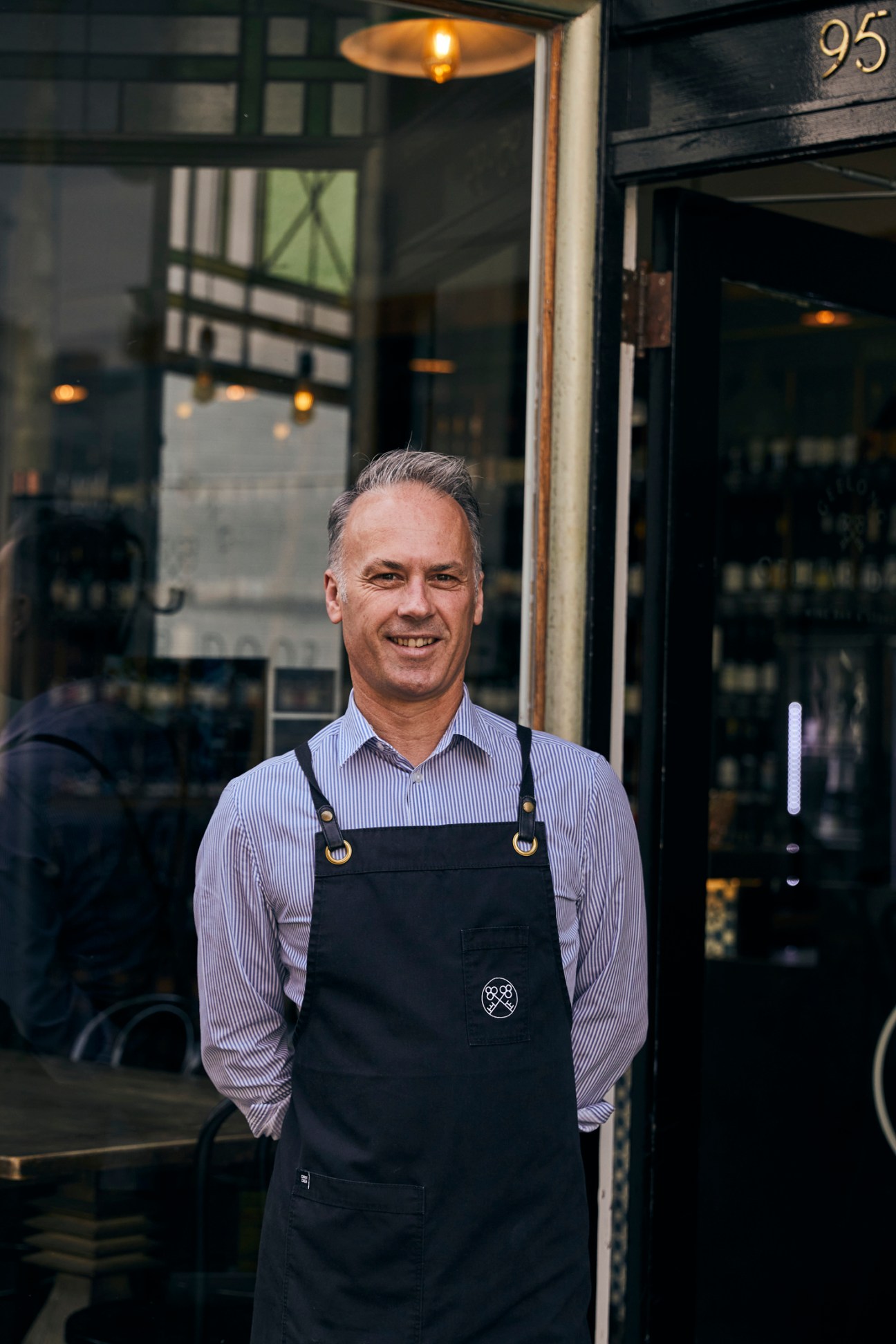 Man in striped shirt and black apron stands smiling outside storefront numbered 95.