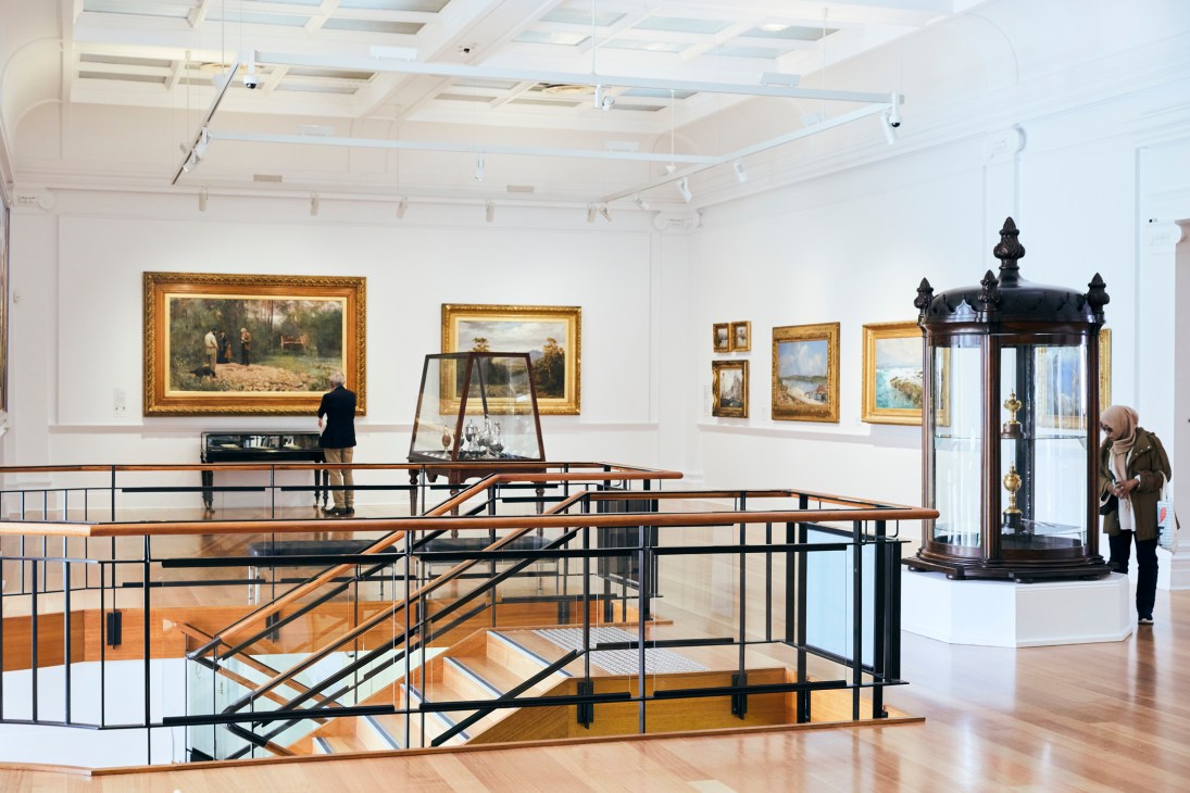 Bright gallery interior with paintings in gilt frames, wooden railings, and visitors viewing artwork.
