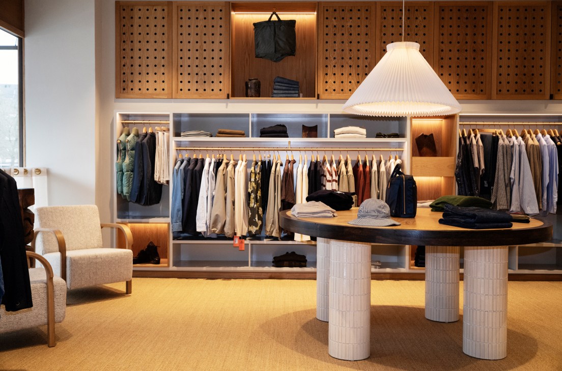 Interior of modern menswear store with wooden perforated wall panels, clothing racks, and central table with pendant lamp