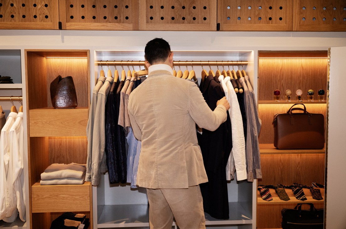 Man in beige sweater browsing clothes in a modern menswear store with wooden shelving and organized displays