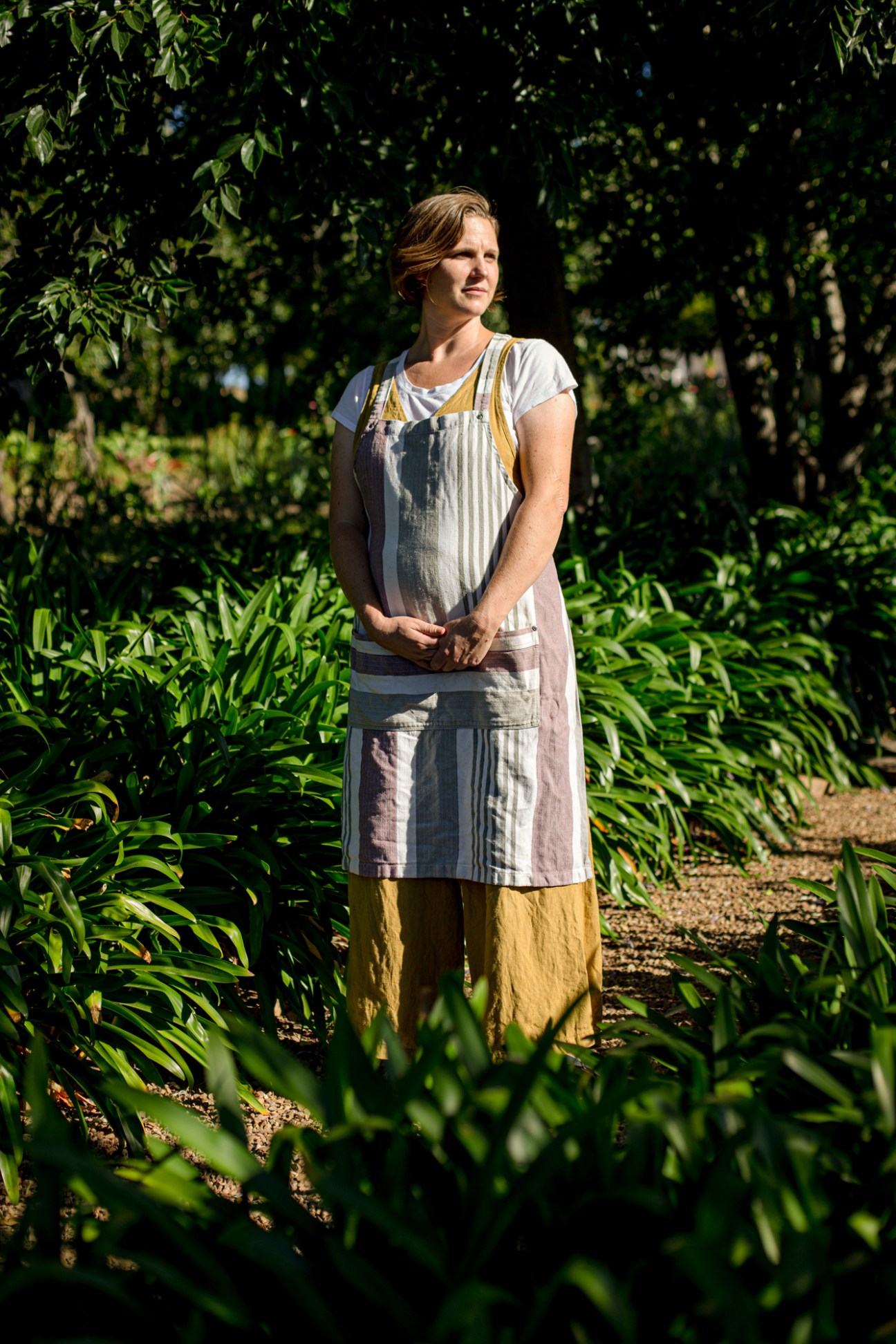Woman in striped apron and yellow pants standing in garden surrounded by lush green plants