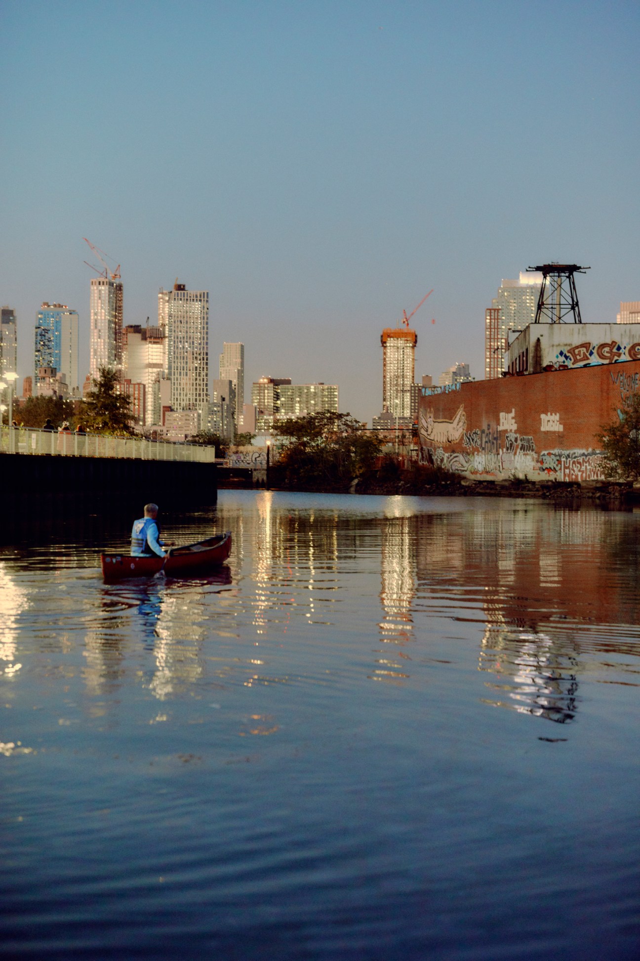 Person paddling red canoe on calm water with city skyline and graffitied buildings in background