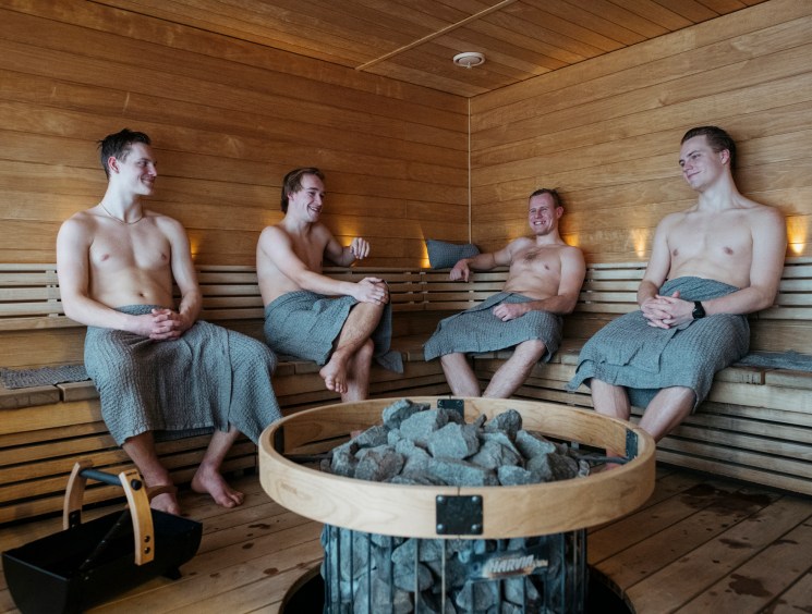 Four men in towels sitting in a wooden sauna with a round stone heater in the center