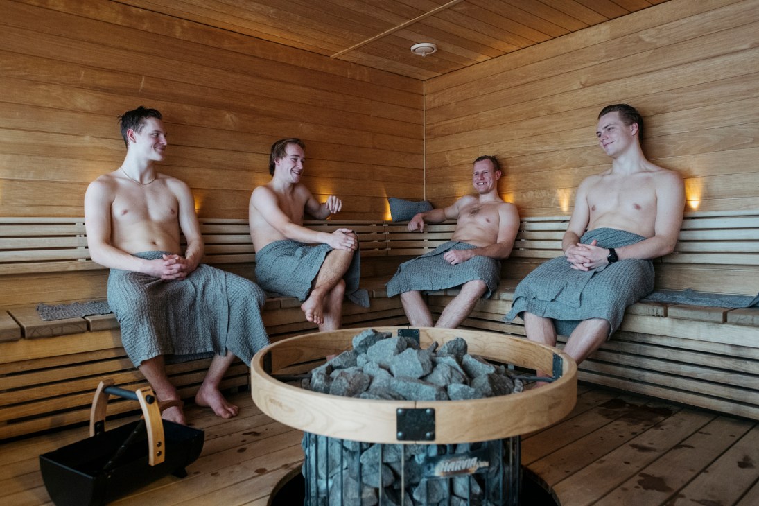Four men in towels sitting in a wooden sauna with a round stone heater in the center
