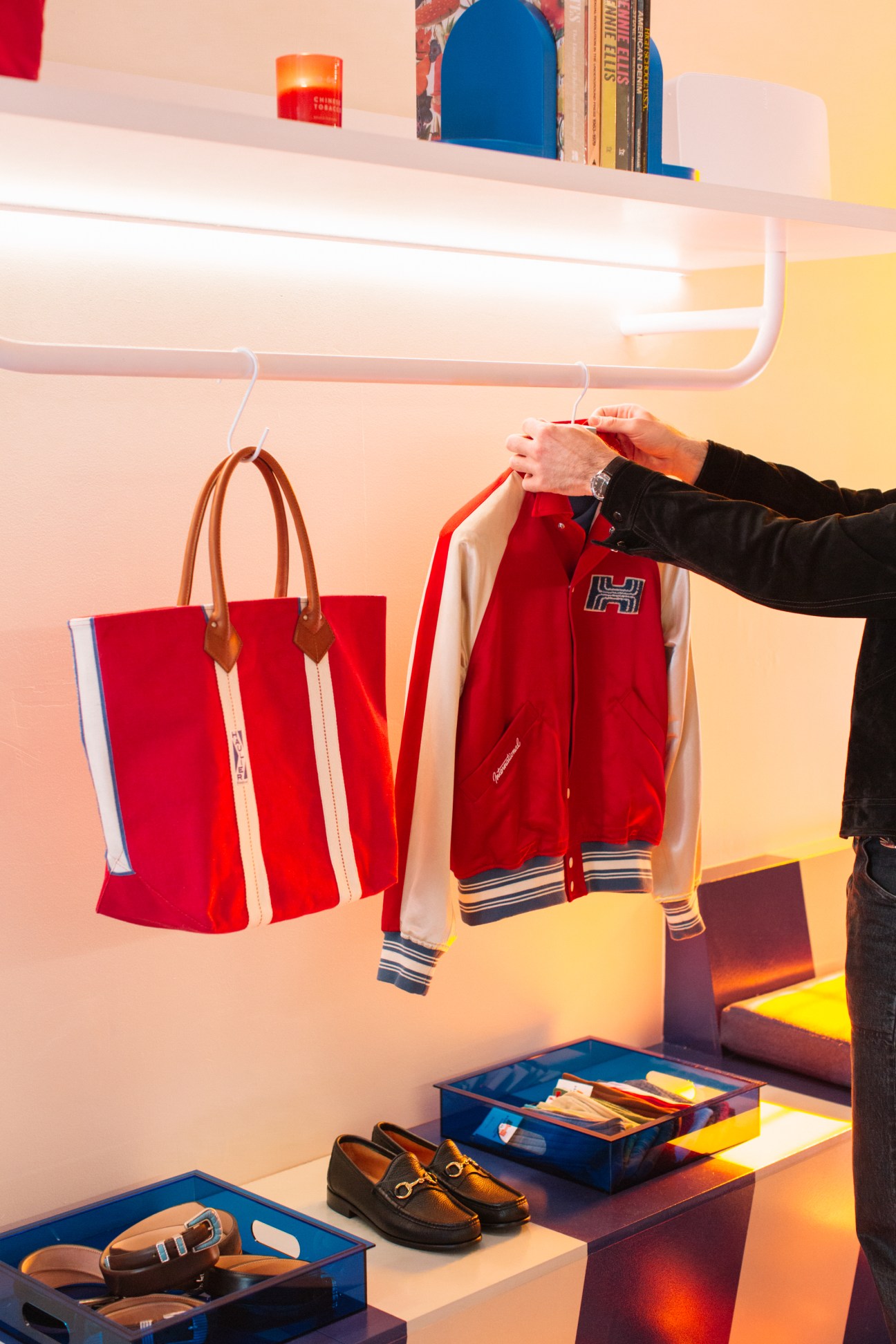 Person reaching for red Haulier varsity jacket hanging on white clothing rack next to red canvas tote bag