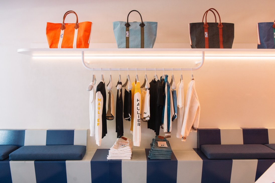 Haulier tote bags in orange, light blue, and black displayed on white shelves with clothing hanging below