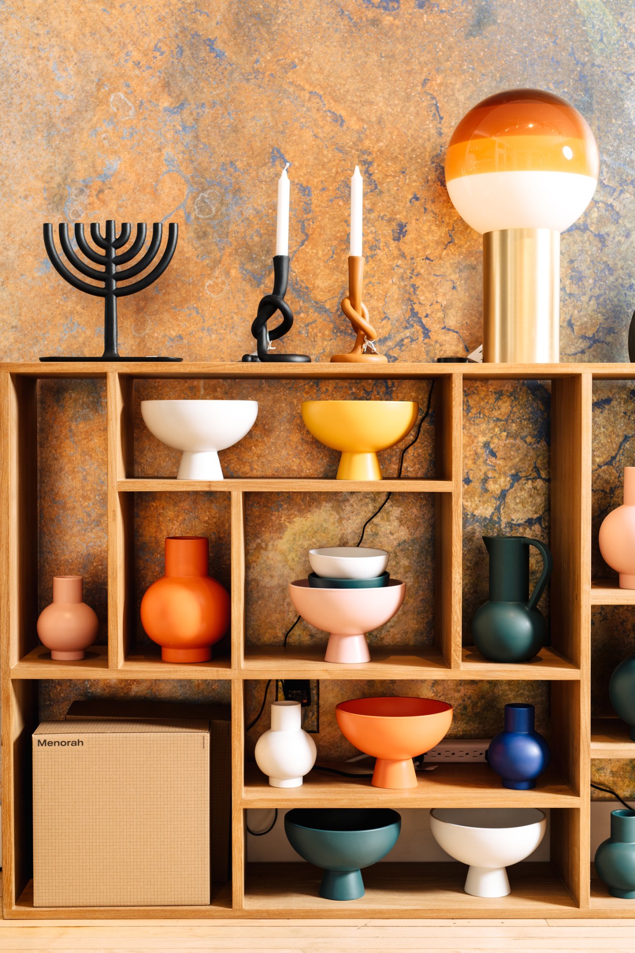Colorful ceramic bowls, vases and home accessories displayed on wooden shelving unit against textured wall