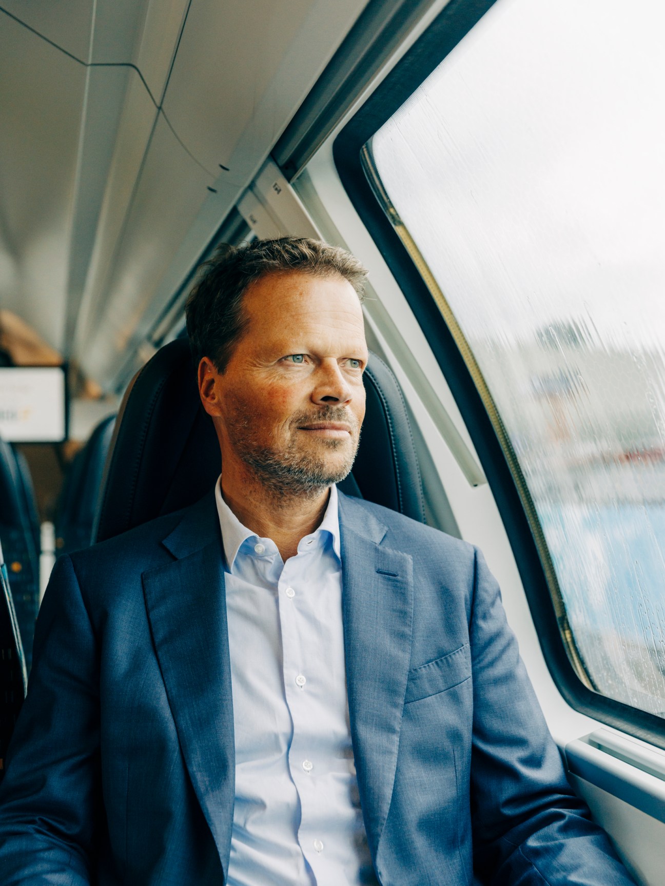Michael Frankenberg in a blue suit sitting by a train window looking thoughtfully outside