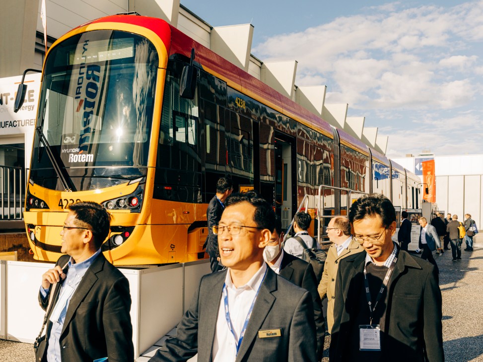 Three men in business attire walking past a modern orange and red train at an indoor exhibition