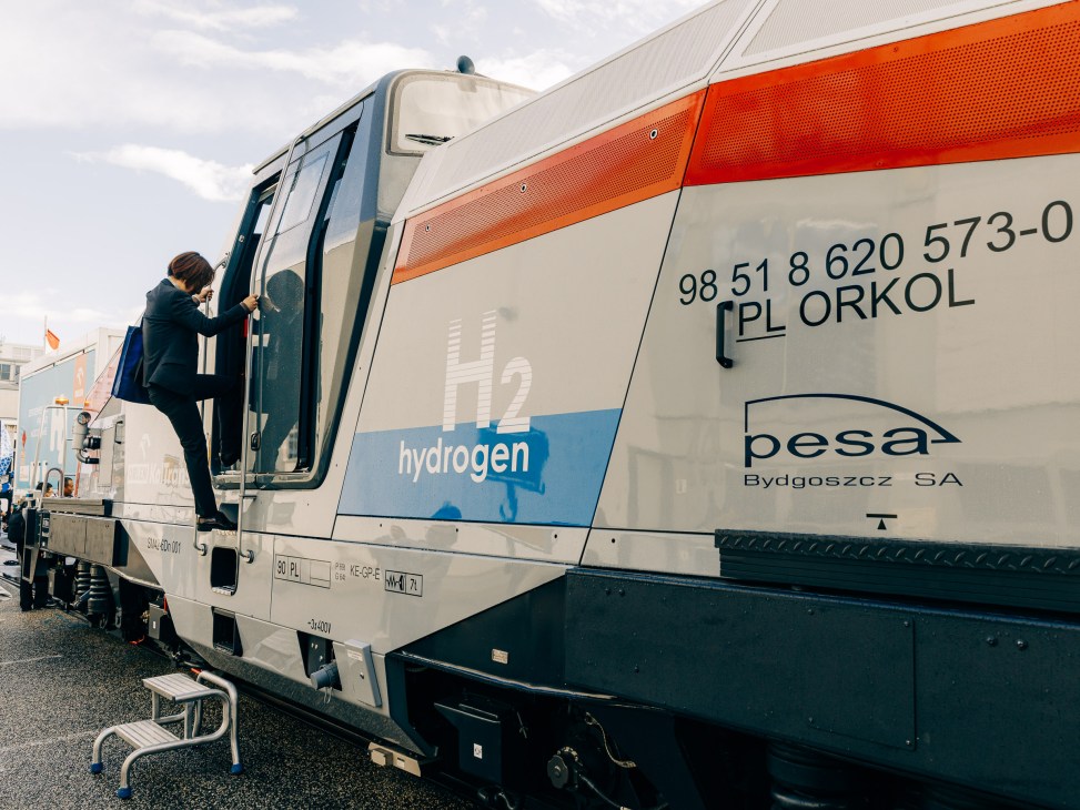 A person in dark clothing boarding Pesa's white and orange hydrogen-powered shunting locomotive at a trade fair