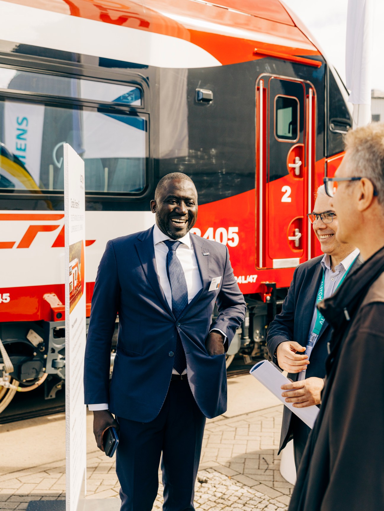 Two men in business suits having a conversation at a trade fair with a red and white train in the background