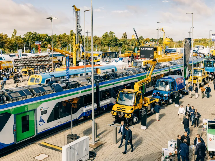 Modern train displayed at outdoor trade fair with yellow maintenance vehicles and visitors walking around