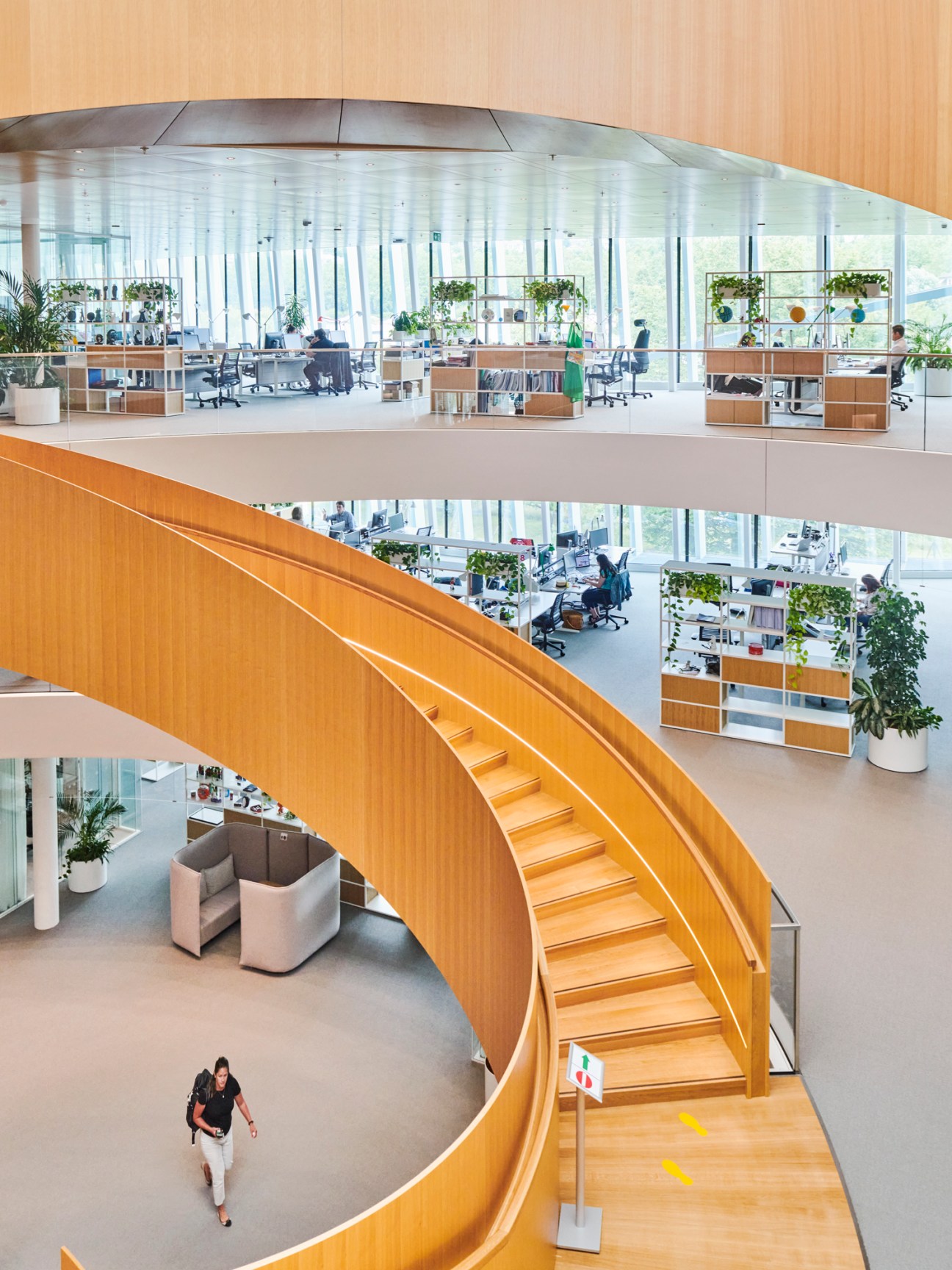 Modern office interior with curved wooden staircase connecting multiple floors, glass walls and open workspace areas