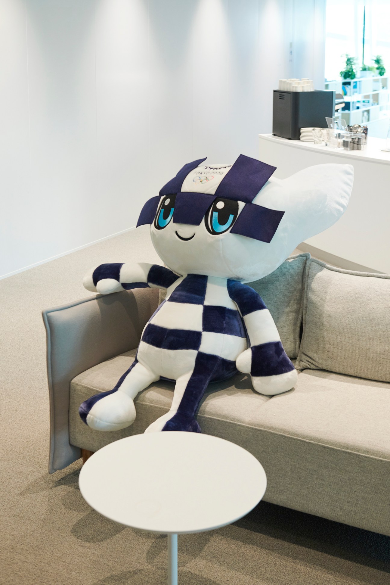 Miraitowa, the Tokyo Olympics mascot, sitting on a modern gray sofa in the IOC headquarters