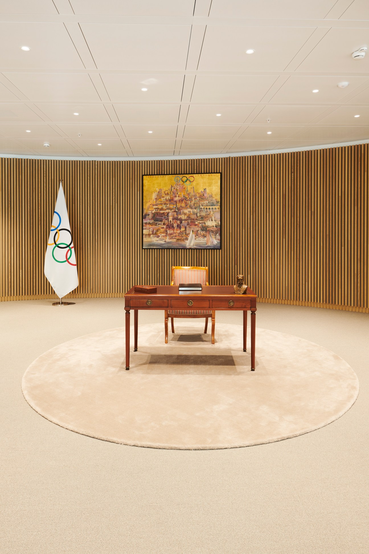 Wooden desk with drawers on circular rug beside Olympic flag in modern office with wood-paneled wall