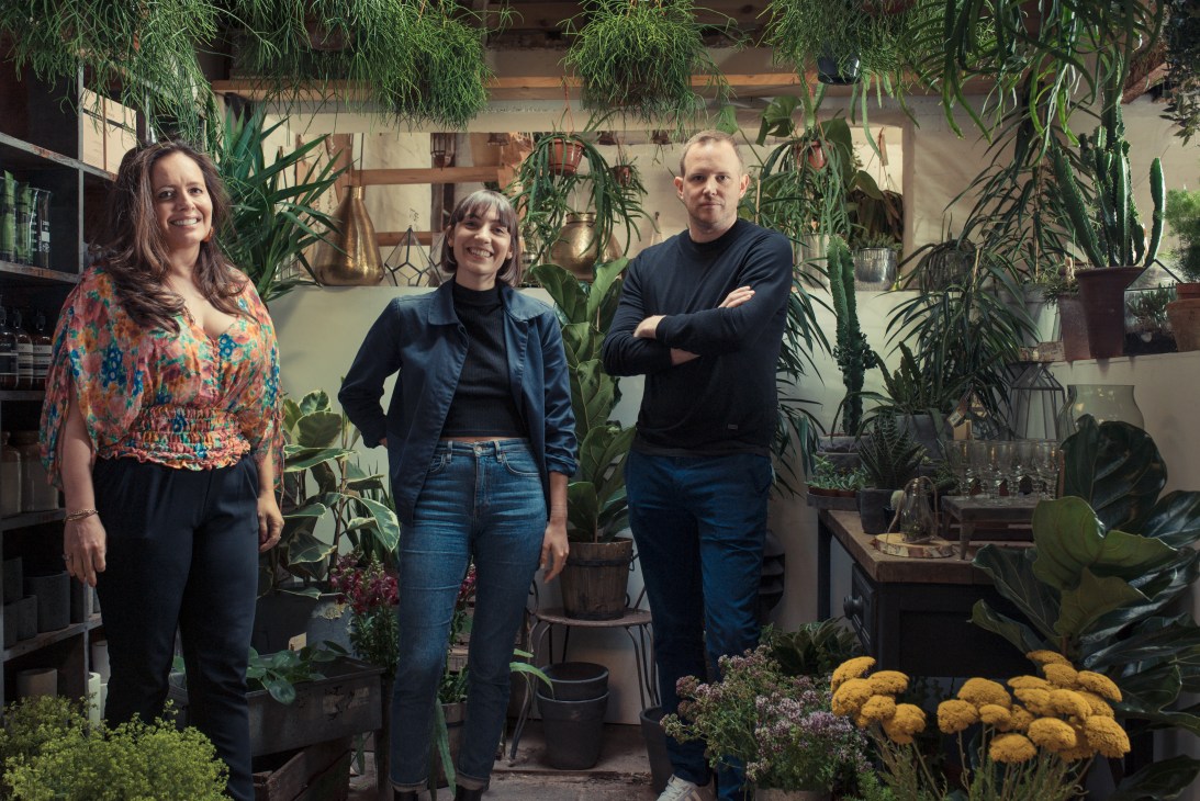 Three people standing in a plant-filled flower shop with hanging greenery and potted plants.