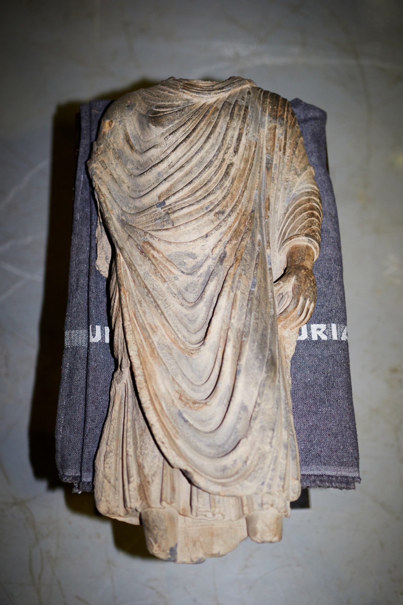 Ancient Buddha torso sculpture in flowing monastic robes displayed on storage shelf