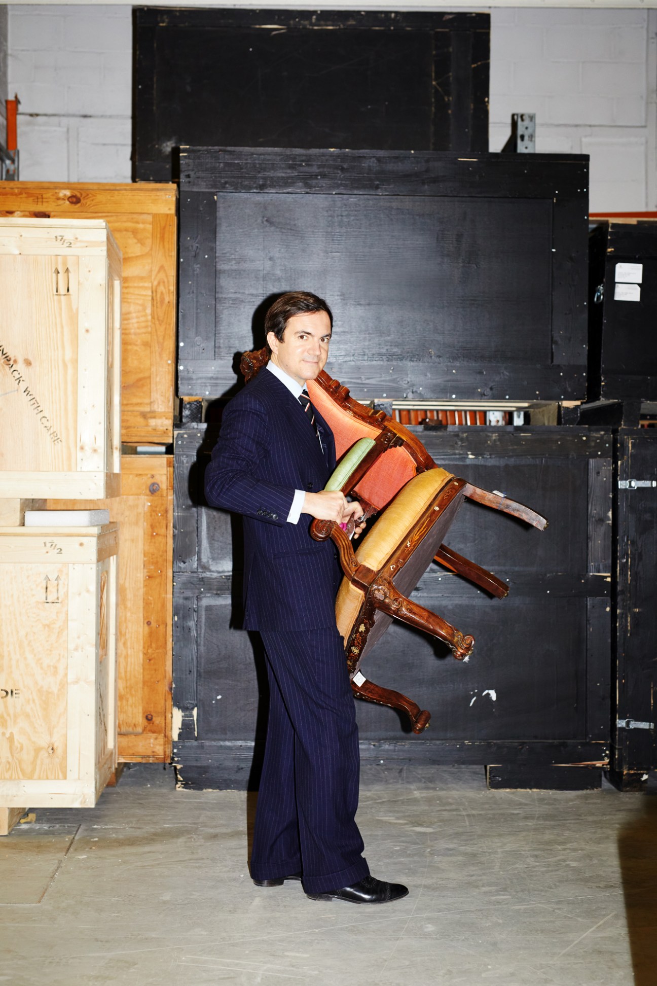 Man in navy pinstripe suit carrying ornate wooden chair in storage facility surrounded by wooden crates