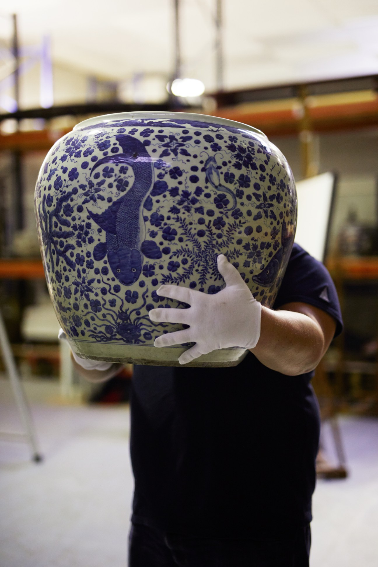 Person wearing white gloves carefully holding a blue and white porcelain urn decorated with aquatic motifs