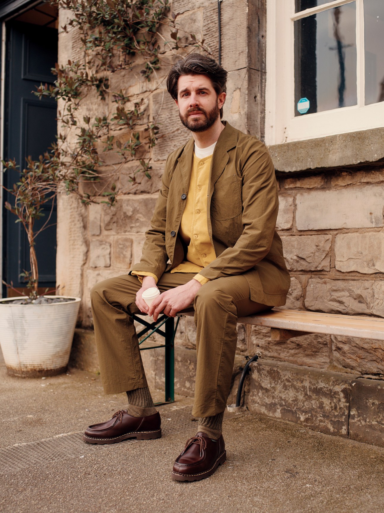 Man in olive green suit and yellow shirt sitting on bench outdoors