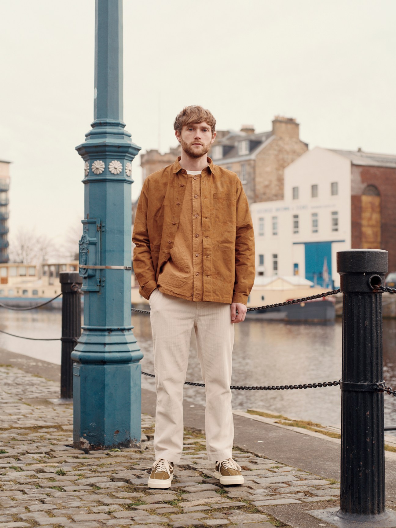 Man in brown corduroy shirt and cream trousers standing by waterfront with historic buildings in background