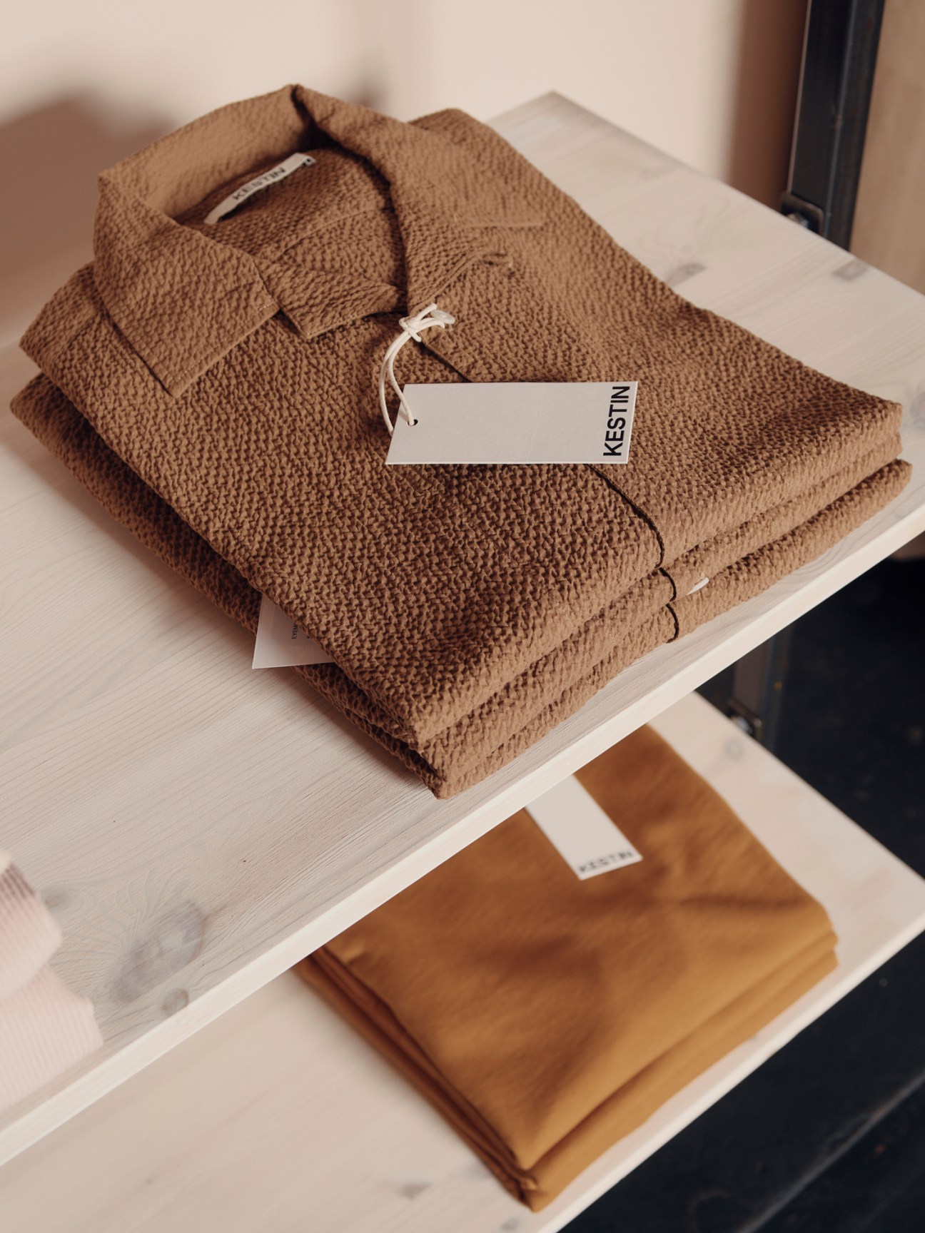 Brown textured shirt with white tag folded on white wooden shelf