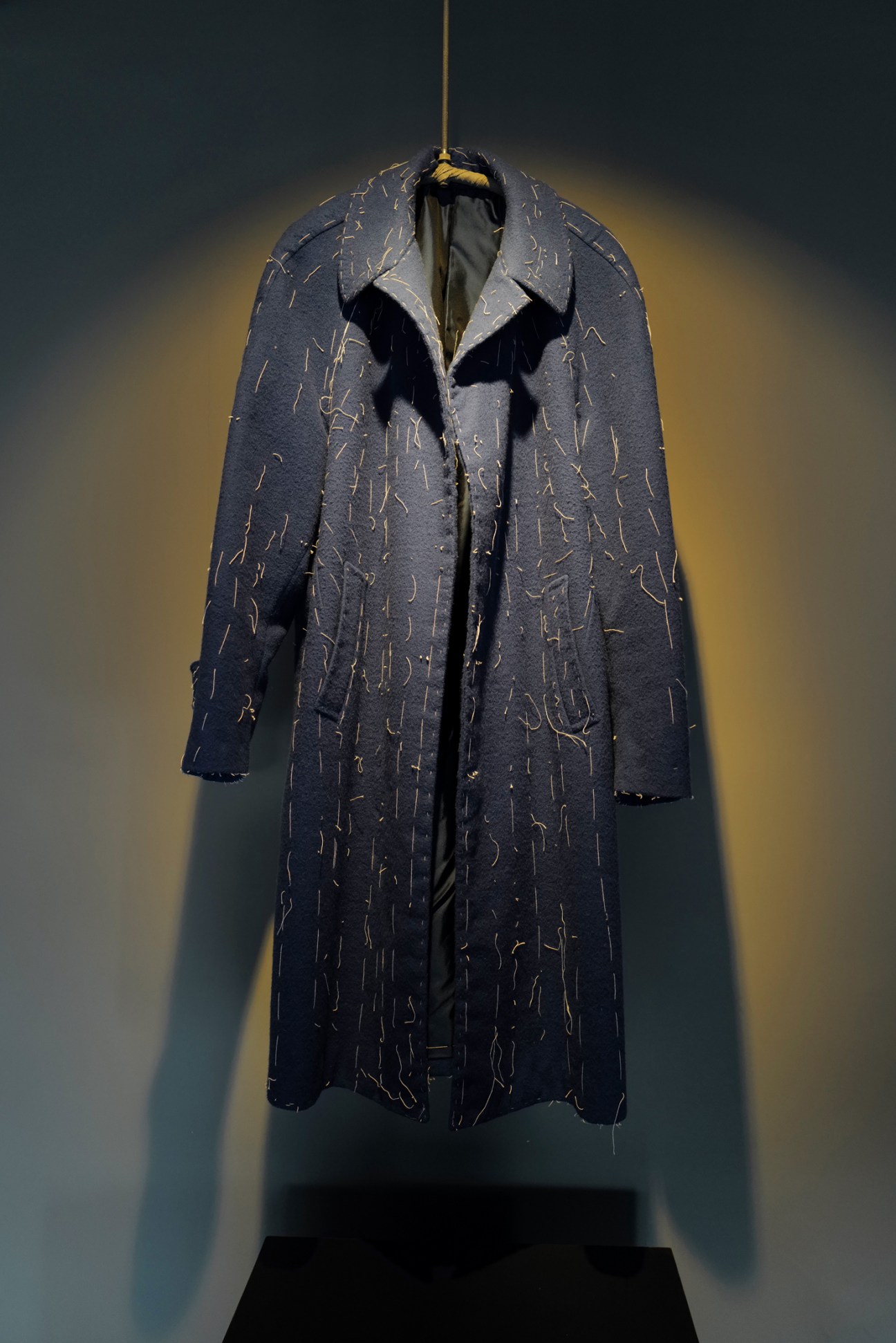 A dark wool coat with detailed stitching displayed on a hanger against a moody lit background